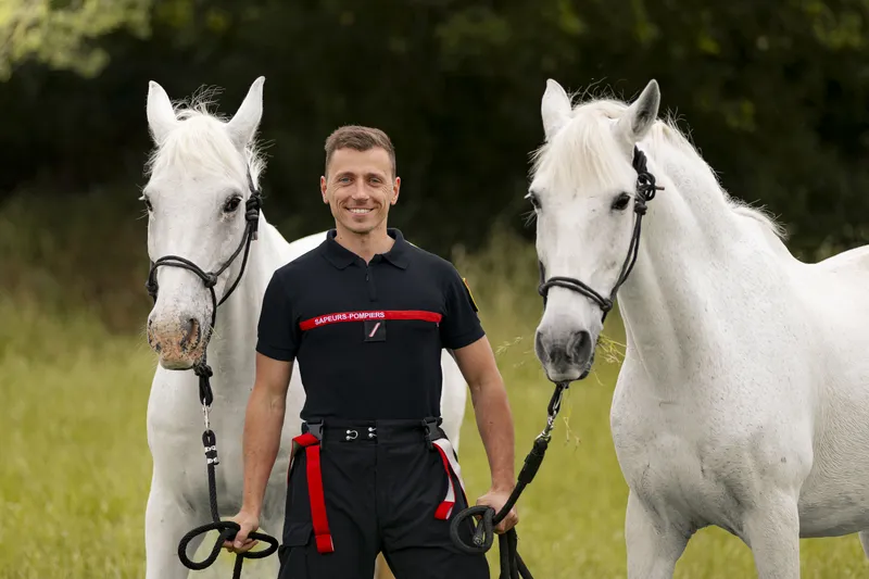 Pompier souriant posant avec deux chevaux blancs dans un pré