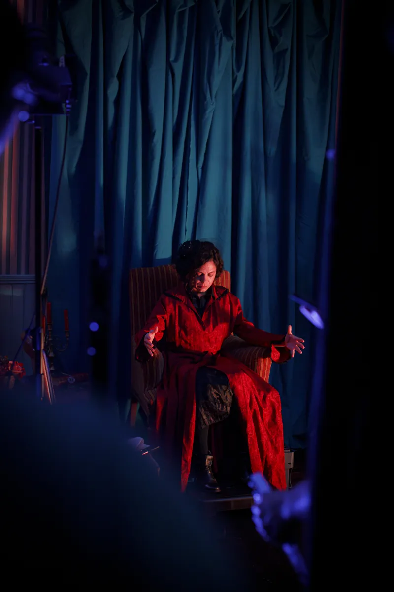 Comédienne en robe rouge assise sur un fauteuil sous un éclairage théâtral bleu