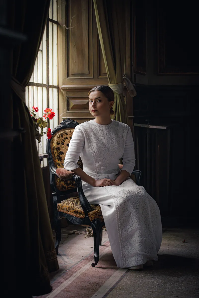 Comédienne en robe blanche assise près d'une fenêtre dans un décor classique