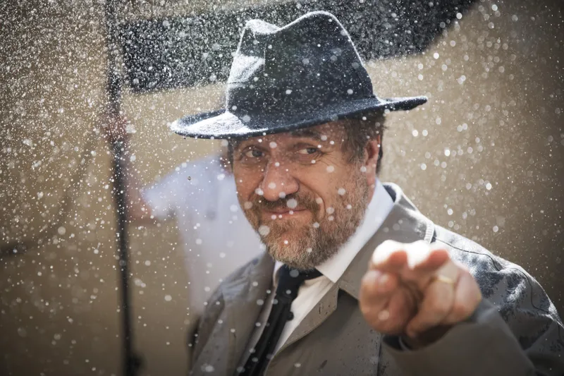 Comédien en costume et chapeau sous la pluie artificielle sur un tournage